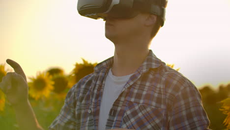 A-male-in-plaid-shirt-and-jeans-uses-VR-glasses-on-the-field-with-sunflowers-for-scientific-article.-These-are-new-technologies-at-sunset.